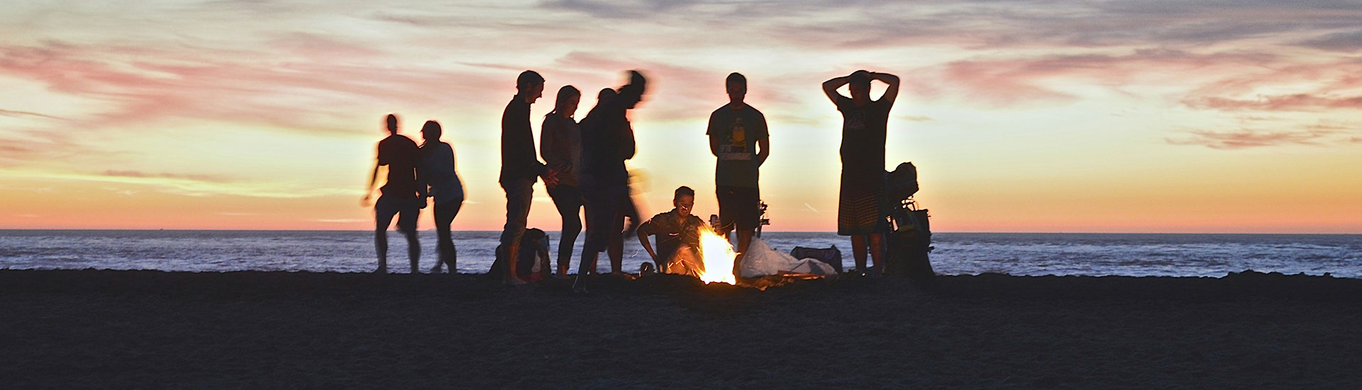 Tio personer står runt en eld, som brinner på en strand. Bakom dem synsa havet och en himmel i solnedgång